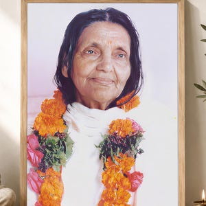 May include: A framed portrait of an elderly person with dark hair, wearing a white garment and a garland of bright orange marigold flowers and pink rose petals. The portrait is encased in a light-colored wooden frame with a simple, clean design.