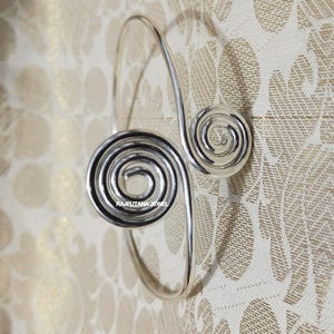 May include: A silver-toned arm cuff with a spiral design. The cuff features two spiral accents, one large and one small, connected by a curved band. The background is a patterned fabric with a floral design. The text "RAJPUTANA JEWEL" is visible.