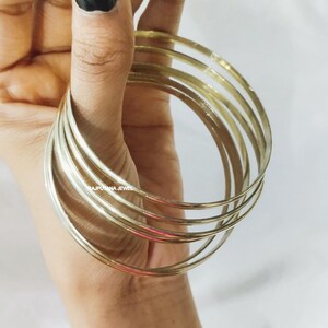 以下が含まれることがあります： 細い金色のバングルが重ねられた様子を、手が持っています。バングルは円形で、金属製のように見えます。背景は無地の白い面です。