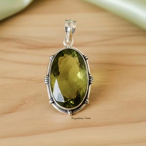 Op de afbeelding: Zilveren hanger met een ovale, olijfgroene edelsteen. De edelsteen is gezet in een gedetailleerde zilveren lijst met decoratieve accenten. De hanger hangt aan een zilveren balein. De tekst "Rajputana Jewel" is zichtbaar.