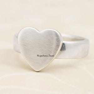 Könnte beinhalten: Ein silberner Ring mit einem herzförmigen Mittelstück. Das Herz hat eine gebürstete Metallstruktur und der Text "Rajputana Jewel" ist sichtbar. Das Band ist breit und glatt und schafft ein schlichtes, aber elegantes Design.