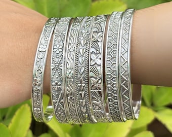 Pode incluir: Um conjunto de pulseiras de prata com intrincados padrões florais e geométricos. As pulseiras são exibidas em um pulso, destacando os designs detalhados. O fundo apresenta folhagem verde.