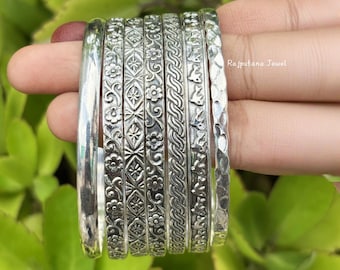 Pode incluir: Um conjunto de pulseiras de prata com vários desenhos em relevo, incluindo padrões florais e geométricos. As pulseiras são exibidas sobre um fundo de folhagem verde. O texto "Rajputana Jewel" é visível.