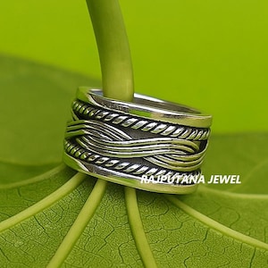 Peut inclure: Une bague en argent avec un motif tissé et des détails en forme de corde. La bague est posée sur un fond de feuille et de tige vertes. Le texte "RAJPUTANA JEWEL" est visible sur la bague.
