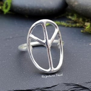 May include: A silver-colored ring featuring a peace symbol design. The ring has a slender band and an openwork peace sign. The ring is displayed on a dark, textured surface with blurred background elements.