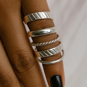 May include: A collection of silver rings in various designs. The rings include a smooth band, a twisted rope design, and rings with diagonal etched lines. The rings are displayed on a hand with black nail polish.