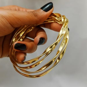 May include: A collection of gold-coloured, twisted metal bangles. The bracelets have a shiny, reflective surface and a textured design. The bangles are stacked together, creating a layered effect. The jewellery is displayed against a neutral background.