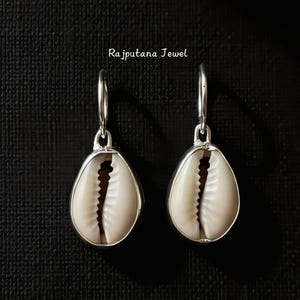 Puede incluir: Un par de pendientes colgantes plateados con conchas de cauri en forma de lágrima. Las conchas son de color blanco crema con un interior marrón oscuro. Los pendientes cuelgan de aros plateados. El texto "Rajputana Jewel" es visible.