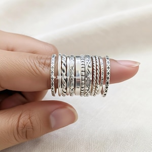May include: A collection of silver rings with various designs, including diamond patterns, twisted rope, and floral motifs. The rings are stacked on a finger, showcasing the different textures and styles. The rings are made of a shiny, metallic material.