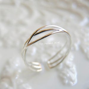 Op de afbeelding: Een zilverkleurige teenring met een gevlochten ontwerp. De ring heeft twee verweven banden die elkaar kruisen, waardoor een open, verstelbaar ontwerp ontstaat. De ring wordt gepresenteerd op een wit oppervlak. De tekst "Rajputana Jewel" is zichtbaar.