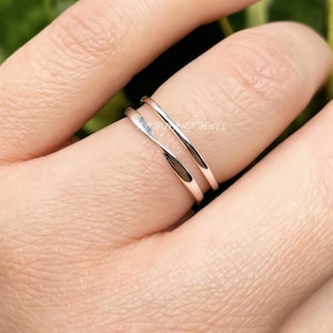 Puede incluir: Primer plano de un anillo plateado con un diseño de doble banda. El anillo se lleva en un dedo, mostrando su estilo elegante y minimalista. La superficie pulida del anillo refleja la luz, resaltando su textura suave. El texto "AJPUANA JEWEL" es visible en el anillo.