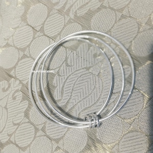 May include: Three silver bangles with a simple, elegant design. The bangles are circular and connected by a small, decorative ring. The jewellery is displayed on a textured, light-coloured surface.