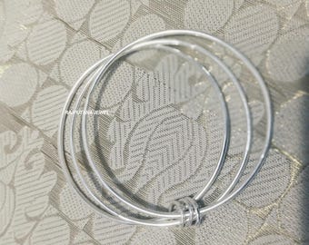 May include: Three silver bangles with a simple, elegant design. The bangles are circular and connected by a small, decorative ring. The jewelry is displayed on a textured, light-colored surface.