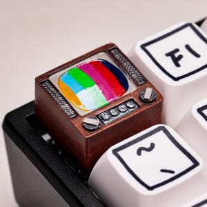 May include: A custom keycap shaped like a vintage television set. The keycap is brown with a colorful screen displaying red, green, blue, and yellow. It is installed on a white keyboard with black lettering and symbols.