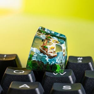 May include: A custom keyboard keycap with a miniature pirate ship scene in clear resin. The ship has a white sail with a skull and crossbones, set against blue water and green islands. The keycap is on black keyboard keys. The keycap is a unique accessory.