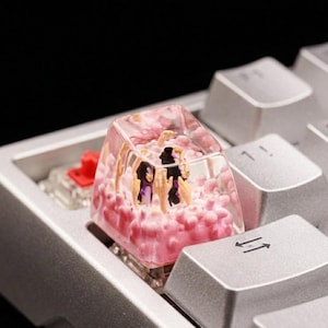 May include: A custom keyboard keycap featuring a clear resin design with a pink, cloud-like interior and miniature figures. The keycap is installed on a silver keyboard, with other keys visible in the background.