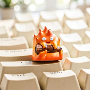 Può includere: Un copritasto artigianale arancione a forma di personaggio di fiamma dei cartoni animati, che tiene un tronco. Il copritasto è posizionato su una tastiera beige. Il personaggio ha una faccia sorridente.