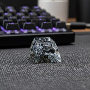 May include: A custom artisan keycap featuring a detailed dragon sculpture encased in clear resin. The keycap is square and sits on a black mechanical keyboard with purple backlighting. The keycap has a gray and brown color scheme.
