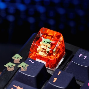 Puede incluir: Una tecla personalizada con una figura verde de Grogu encerrada en un bloque de resina transparente con un interior rojo. La tecla está en un teclado negro, con tres pegatinas de Grogu cerca.