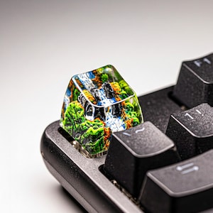 May include: A custom keyboard keycap featuring a miniature landscape with green trees, a waterfall, and blue sky. The keycap is transparent, allowing the landscape to be visible. The keycap is installed on a black keyboard.