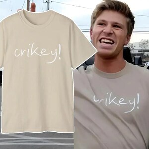 May include: A beige t-shirt with the word "crikey!" in white script. The shirt is displayed on a hanger and is also being worn by a person. The person is smiling and appears to be holding the shirt.