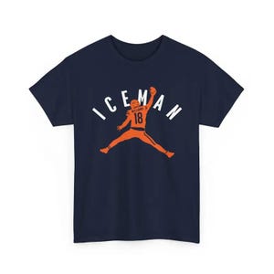 May include: Navy blue t-shirt featuring an orange silhouette of a football player in mid-air, with the word "ICEMAN" above. The player's jersey displays the number 18. A stylish graphic tee.