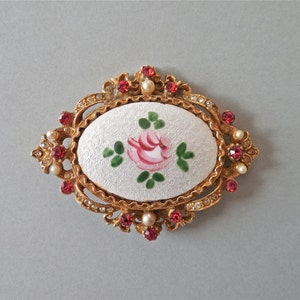 Puede incluir: Un broche de tono dorado con un camafeo ovalado blanco que presenta una rosa rosa pintada a mano con hojas verdes. El camafeo est&aacute; rodeado por un borde de piedras de imitaci&oacute;n y piedras falsas rojas.