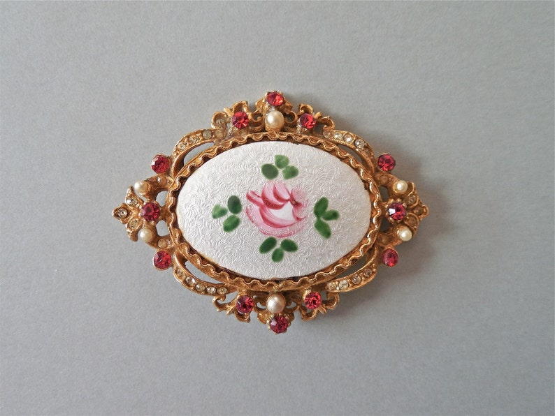 Puede incluir: Un broche de tono dorado con un camafeo ovalado blanco que presenta una rosa rosa pintada a mano con hojas verdes. El camafeo est&aacute; rodeado por un borde de piedras de imitaci&oacute;n y piedras falsas rojas.