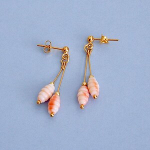May include: A pair of gold-colored dangle earrings with a post back. Each earring features two strands with a light pink and orange-toned, oval-shaped bead. The earrings are set against a light blue background.