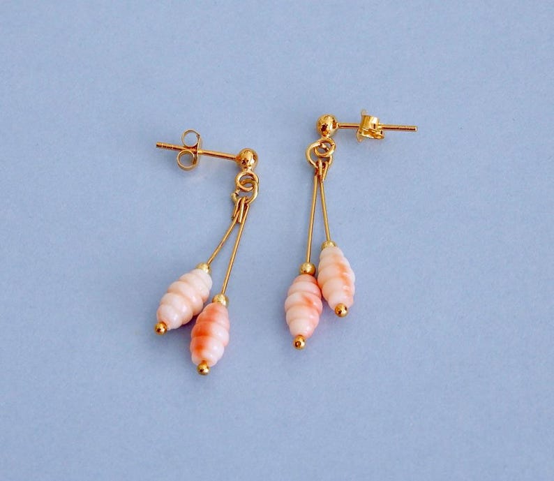 May include: A pair of gold-colored dangle earrings with a post back. Each earring features two strands with a light pink and orange-toned, oval-shaped bead. The earrings are set against a light blue background.