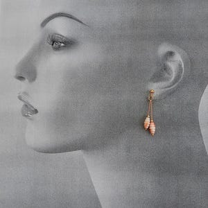 May include: A pair of gold-colored drop earrings with two coral-colored, oblong beads suspended from each earring. The earrings are displayed against a grayscale background of a person's face.