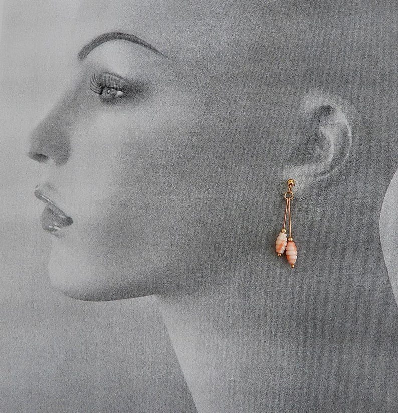 May include: A pair of gold-colored drop earrings with two coral-colored, oblong beads suspended from each earring. The earrings are displayed against a grayscale background of a person's face.
