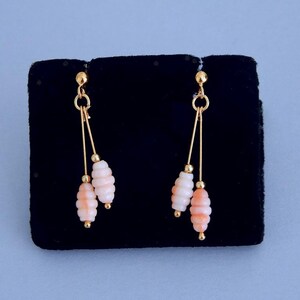 May include: Dangling earrings with a gold-tone metal design. Each earring features a gold stud, two gold wires, and two coral-colored, stacked bead clusters. The earrings are displayed on a black velvet surface against a light blue background.