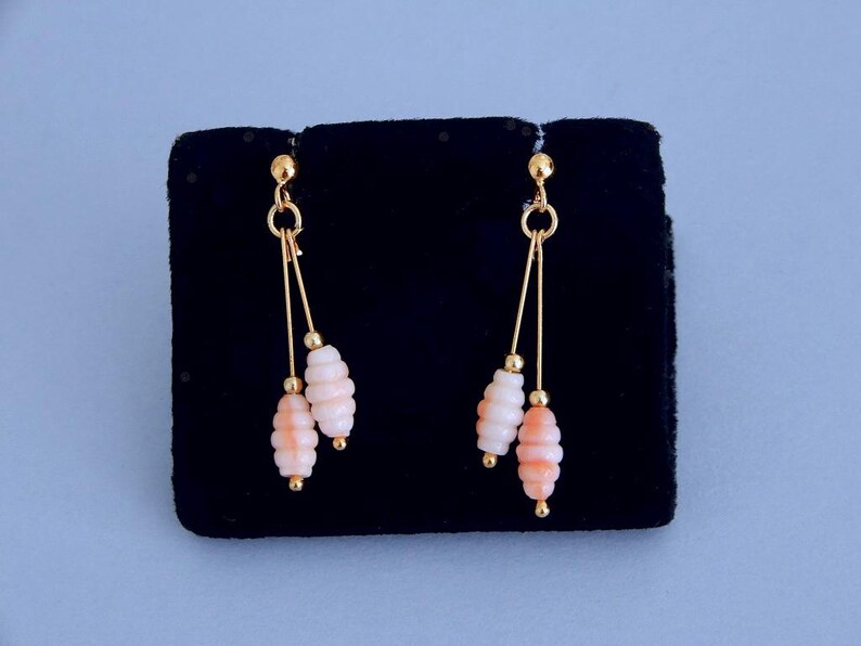 May include: Dangling earrings with a gold-tone metal design. Each earring features a gold stud, two gold wires, and two coral-colored, stacked bead clusters. The earrings are displayed on a black velvet surface against a light blue background.