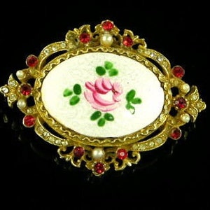 Puede incluir: Un broche de tono dorado con un camafeo ovalado blanco que presenta una rosa rosa pintada a mano con hojas verdes. El camafeo est&aacute; rodeado por un marco decorativo con diamantes de imitaci&oacute;n rojos y transparentes.