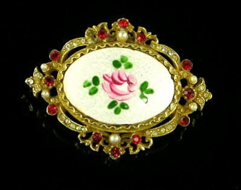 Puede incluir: Un broche de tono dorado con un camafeo ovalado blanco que presenta una rosa rosa pintada a mano con hojas verdes. El camafeo est&aacute; rodeado por un marco decorativo con diamantes de imitaci&oacute;n rojos y transparentes.