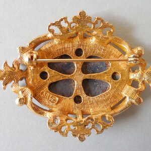 Puede incluir: Broche dorada con un dise&ntilde;o ornamentado y un centro azul oscuro. La parte posterior de la broche tiene la palabra "ARTCO" estampada.