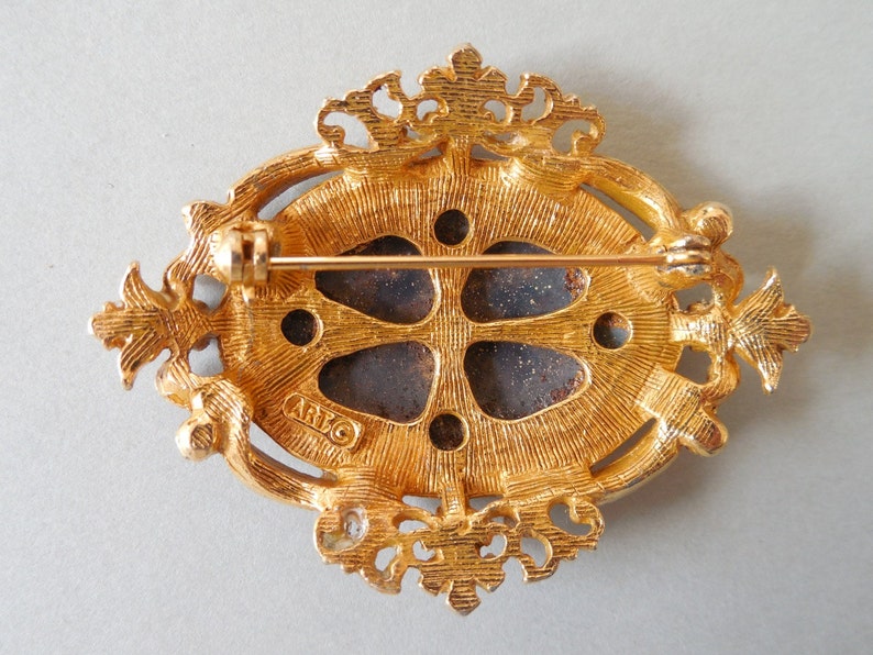 Puede incluir: Broche dorada con un dise&ntilde;o ornamentado y un centro azul oscuro. La parte posterior de la broche tiene la palabra "ARTCO" estampada.