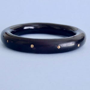 May include: A smooth, dark brown, circular bangle bracelet with a polished finish. The exterior surface of the bangle is evenly decorated with small, round, metallic gold-colored studs spaced at regular intervals around the entire circumference.