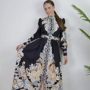 May include: A long black dress with a high collar and long sleeves. The dress features a paisley pattern in shades of blue, gold, and white. A matching belt cinches the waist. The dress is buttoned down the front and has a slit in the skirt.