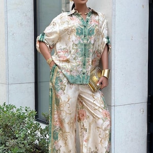 May include: A matching floral print silk shirt and wide-leg pants set in cream, green, and pink. The shirt has a button-down front and rolled-up sleeves. The pants have a green stripe down the side. A gold clutch and bracelet complete the look.