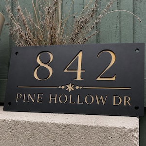 May include: Black rectangular house sign displaying the numbers "842" in a light tan hue. The text "PINE HOLLOW DR" is below the numbers. The sign features a floral design and is mounted on a concrete surface.