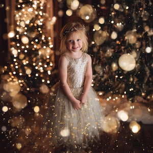 May include: A young girl in a gold sequined dress and a delicate headpiece smiles in front of a Christmas tree with twinkling lights. The dress has a tulle skirt with gold confetti. The background is softly blurred with bokeh.