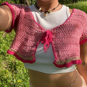 Könnte beinhalten: Ein rosa Häkel-Bolero mit kurzen Ärmeln und einem Bindverschluss. Die Kanten sind mit einem dunkleren Rosa und einem Hauch von Grün verziert. Der Bolero wird über einem weißen Top getragen.
