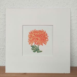 Puede incluir: Impresión enmarcada de una vibrante flor de crisantemo naranja con hojas verdes. La obra de arte está centrada en un fondo blanco dentro de un passepartout blanco, contra una pared neutra. Se muestra sobre una superficie de madera.