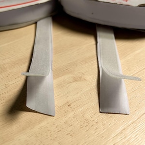 May include: Close-up of two gray hook-and-loop fastener straps, with the hook side exposed. The straps are attached to a gray object, possibly footwear. The background is a light brown wooden surface.