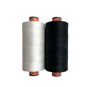 Può includere: Due rocchetti di filo, uno bianco e uno nero, sono esposti su sfondo bianco. Il rocchetto bianco ha la scritta "1000 m" e il rocchetto nero ha la scritta "AMANN". I rocchetti sono cilindrici e sembrano essere fatti di filo ben avvolto.