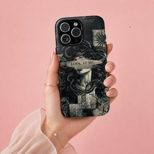Peut inclure: Une coque de smartphone noire et blanche présentant une illustration détaillée d'une figure aux cheveux de serpents, rappelant Méduse. Un graphisme en forme de coup de pinceau horizontal sur les yeux contient le texte LOOK AT ME en gras.