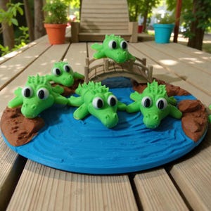 Peut inclure: Un diorama fantaisiste fait à la main avec cinq figurines d'alligators verts aux grands yeux cerclés de blanc. Ils sont disposés autour d'un plan d'eau bleu avec un petit pont et des masses terrestres brunes. La base est de forme circulaire.