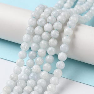 May include: Close-up of strands of light blue-grey gemstone beads. The beads are round and have a slightly frosted appearance. The beads are strung together with a light blue string. The background is white and light blue.